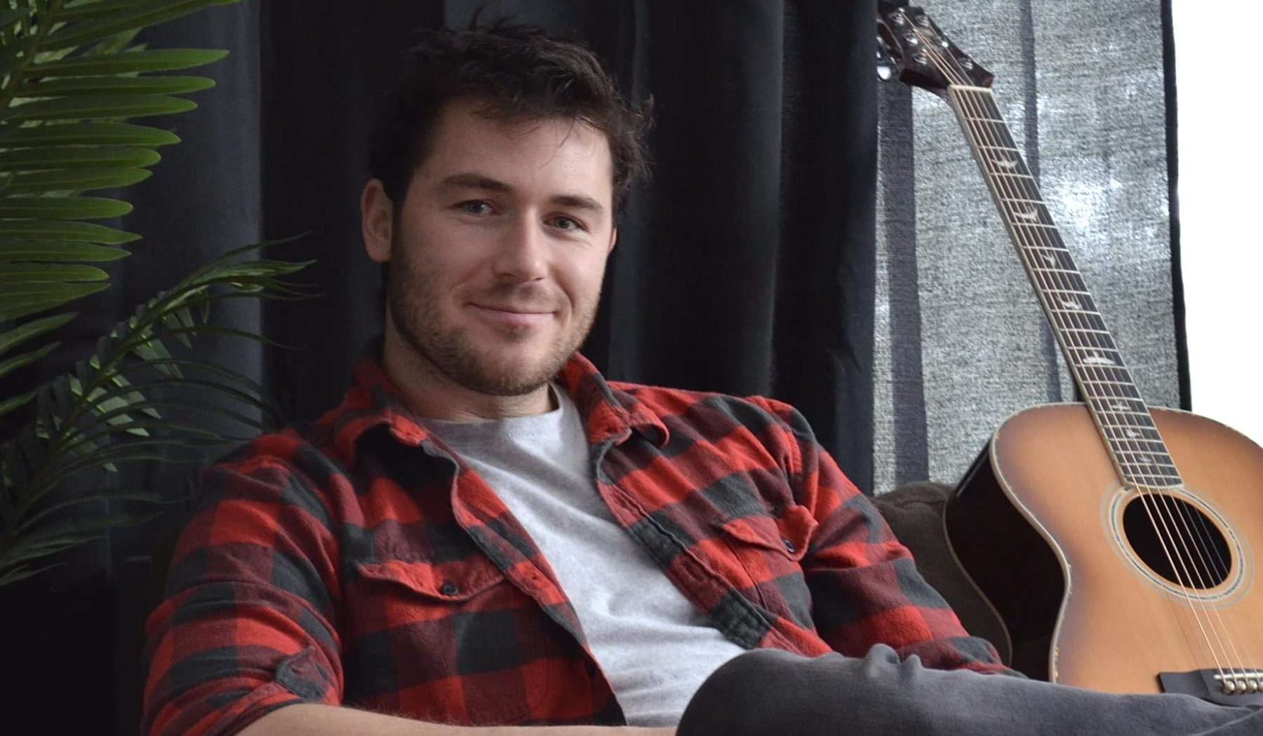 Nathan couture assis dans une chaise dans un salon, sa guitare en fond à côté de lui.