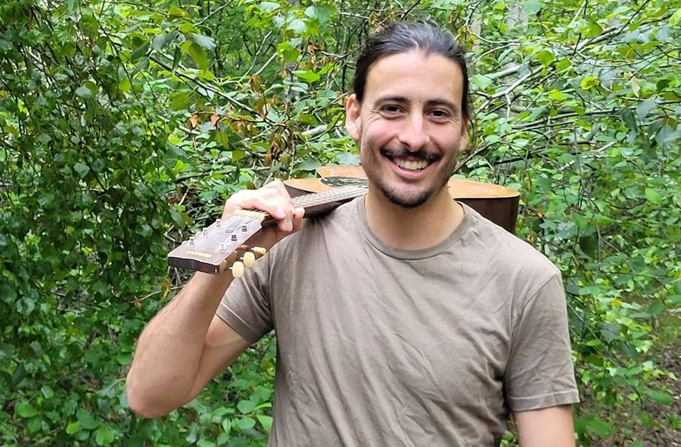 Félix Leroux tenant sa guitare sur son épaule en souriant. Les feuilles d'un arbres sont visibles derrière lui.